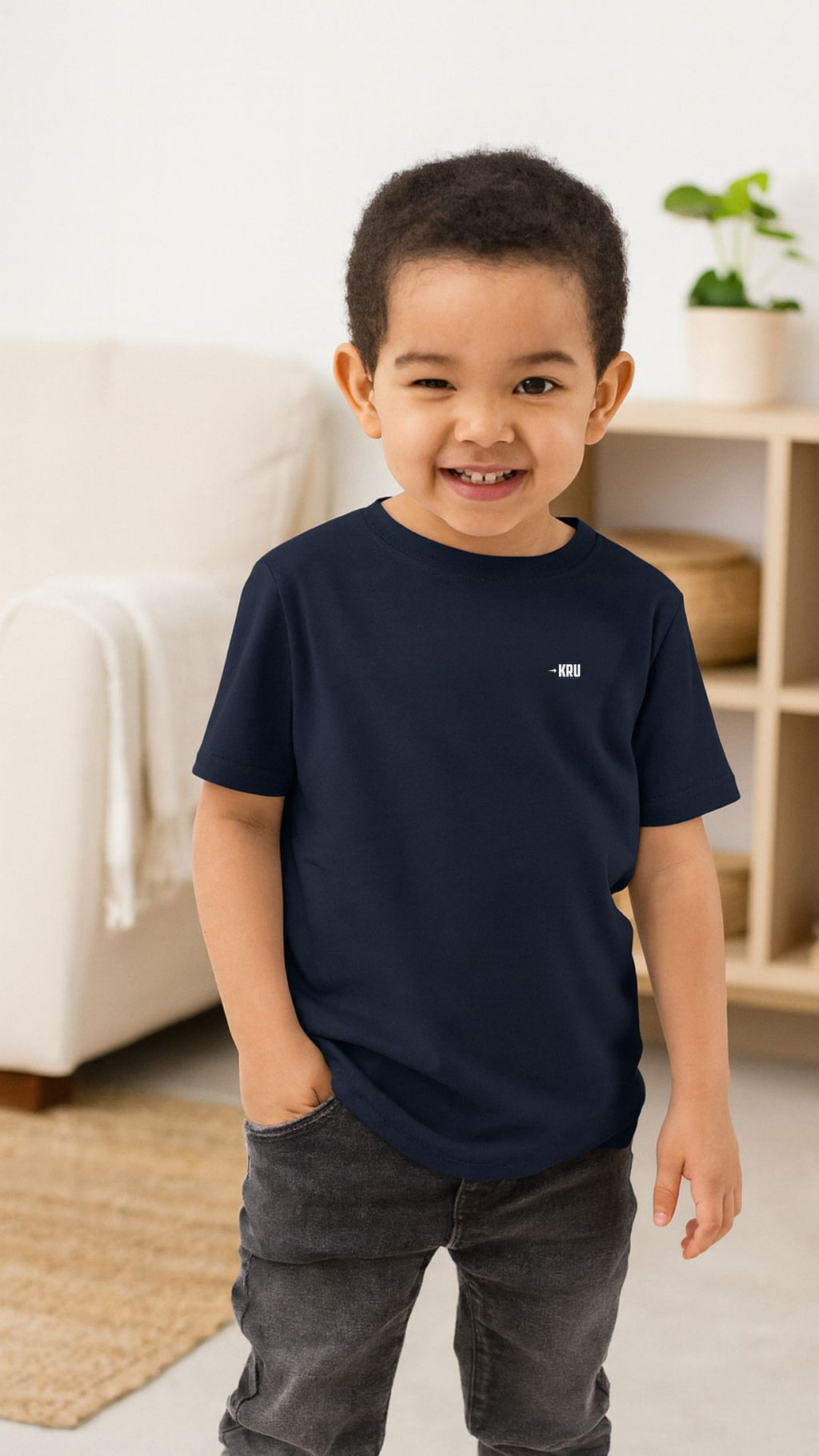 Child wearing a navy blue t-shirt with a small logo, standing in a room with a white couch and plant in the background.