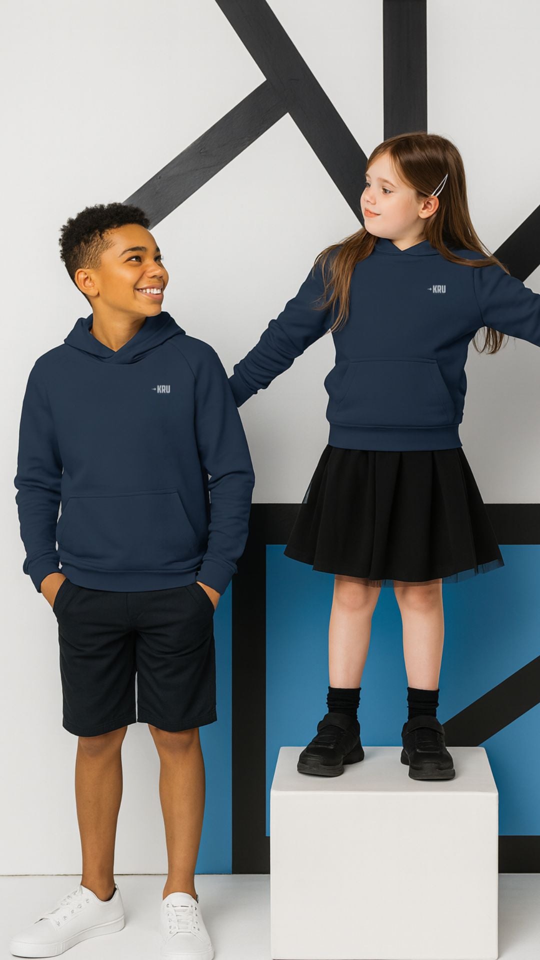 Two children wearing navy blue hoodies and black shorts in front of a geometric wall design.